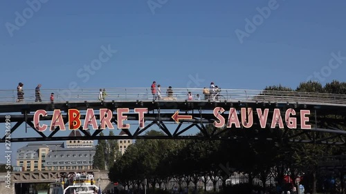 Paris, France - April 5, 2019: Parisian boat passing under a bridge at the Parc de la Villette with the inscription Cabaret Sauvage. People crossing the bridge. Sunny summer with blue sky. Tight shot. photo