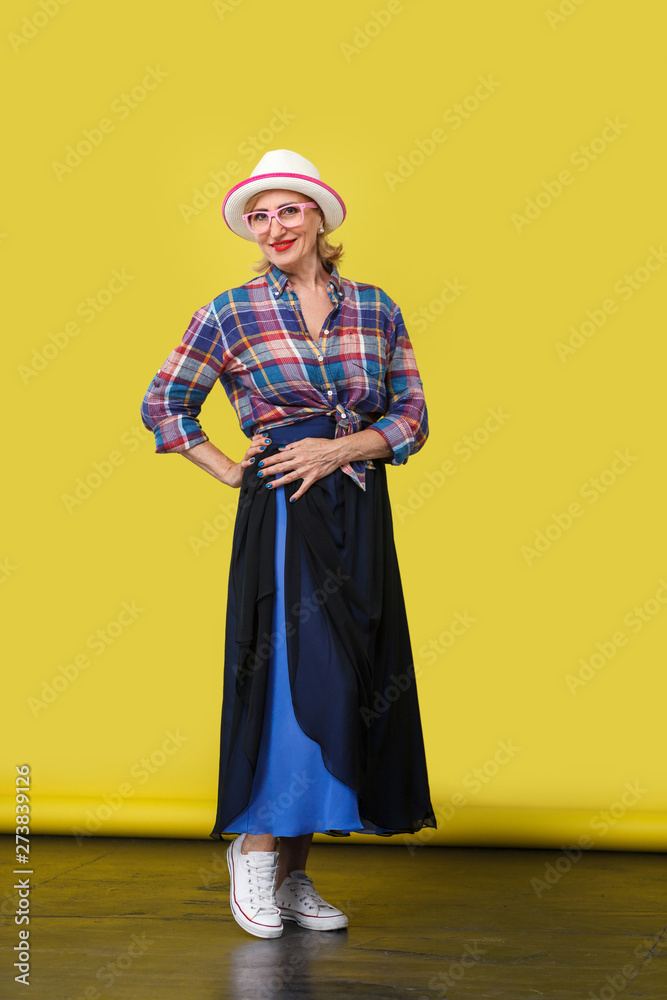 full length portrait of beautiful happy modern stylish mature woman in casual style with hat and eyeglasses standing, posing and looking at camera. indoor studio shot on yellow wall background. full length portrait of beautiful happy modern stylish mature woman in casual style with hat and eyeglasses standing, posing and looking at camera. indoor studio shot on yellow wall background.