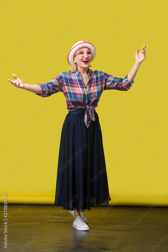 full length portrait of happy modern stylish mature woman in casual style with hat and eyeglasses standing with raised arms and surprised face looking. indoor studio shot on yellow wall background.