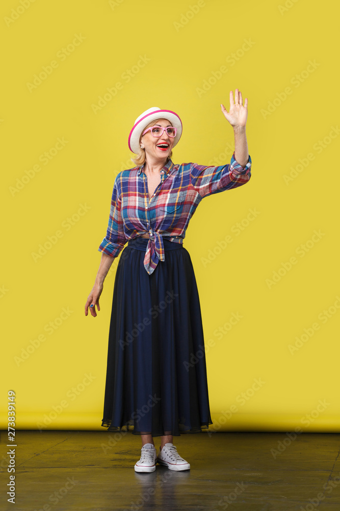 full length portrait of excited modern stylish mature woman in casual style with hat, eyeglasses standing, waving her hand and looking with toothy smile. indoor studio shot on yellow wall background.