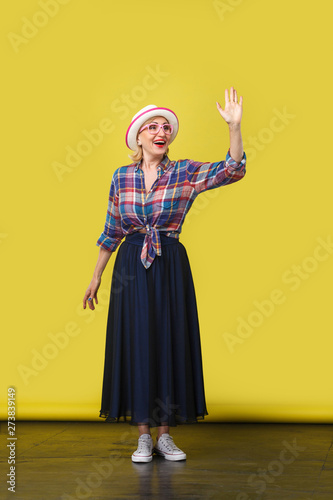 full length portrait of excited modern stylish mature woman in casual style with hat, eyeglasses standing, waving her hand and looking with toothy smile. indoor studio shot on yellow wall background.