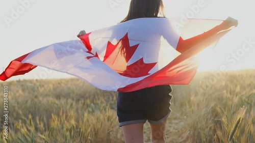 Young happy canadian girl running with open arms over wheat field and holding Canada flag at sunset. Slow motion photo