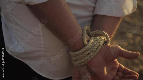 Hands of man tied in the back by a thick rope in the Moroccan desert in the middle of a heat wave during the sunny summer. Man standing still, kidnapped by criminals. Sun rays on the hands. Close up.