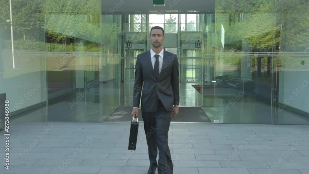 Handsome caucasian businessman wearing a suit coming out of work going through a transparent sliding door entrance to bay window with nature reflections, financial building in the business center.