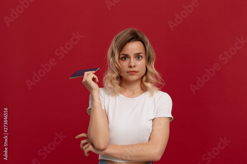 Portrait of a beautiful girl with curly blond hair dressed in a white t-shirt standing on a red background. Model holds passport of blue color and looks at the camera.