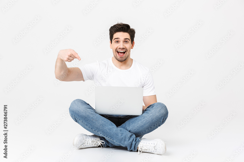 Happy excited young man posing isolated over white wall using laptop computer pointing sitting on floor. Happy excited young man posing isolated over white wall using laptop computer pointing sitting on floor.