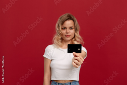 Portrait of a girl with curly blond hair in a white t-shirt standing on a red background. Model smiles at the camera and holds bank card of black color.