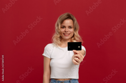 Portrait of a girl with curly blond hair in a white t-shirt standing on a red background. Model smiles at the camera and holds bank card of black color.