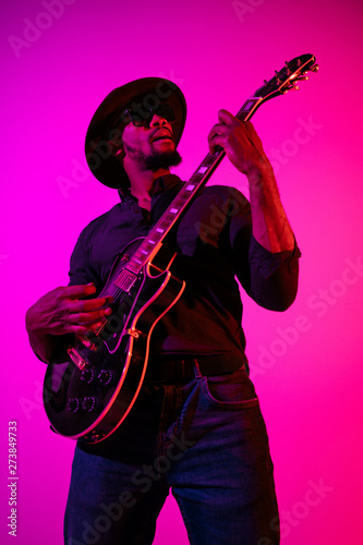 Young african-american musician playing the guitar like a rockstar on gradient purple-pink background in neon light. Concept of music, hobby. Joyful attractive guy improvising and singing a song.