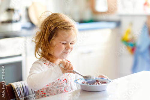 Adorable toddler girl eating healthy cereal with milk for breakfast. Cute happy baby child in colorful clothes sitting in kitchen and having fun with preparing oats  cereals. Indoors at home