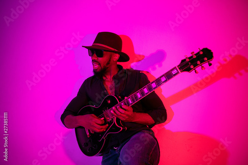 Young african-american musician playing the guitar like a rockstar on gradient purple-pink background in neon light. Concept of music, hobby. Joyful attractive guy improvising. Colorful portrait. © master1305