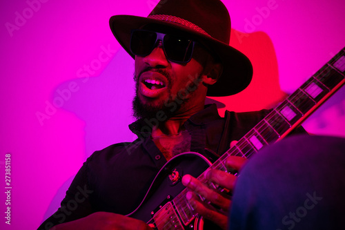 Young african-american musician playing the guitar like a rockstar on gradient purple-pink background in neon light. Concept of music, hobby. Joyful attractive guy improvising. Colorful portrait.