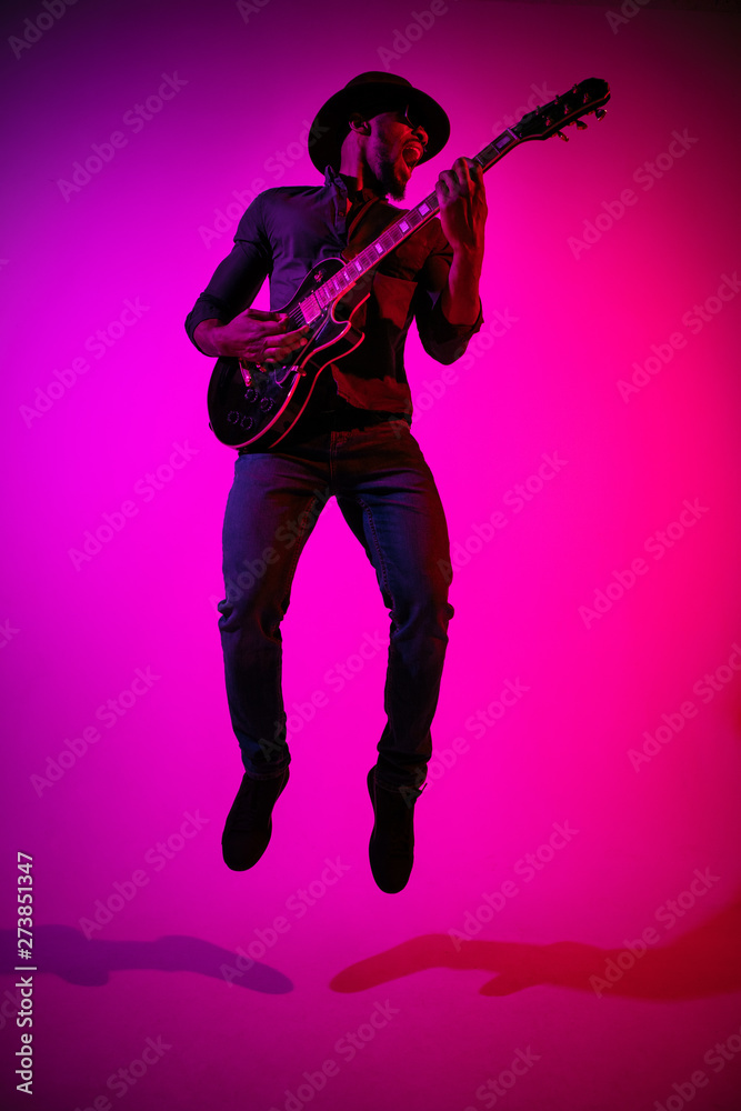 Young african-american musician playing the guitar like a rockstar on gradient purple-pink background in neon light. Concept of music, hobby. Joyful attractive guy improvising. Colorful portrait. Young african-american musician playing the guitar like a rockstar on gradient purple-pink background in neon light. Concept of music, hobby. Joyful attractive guy improvising. Colorful portrait.