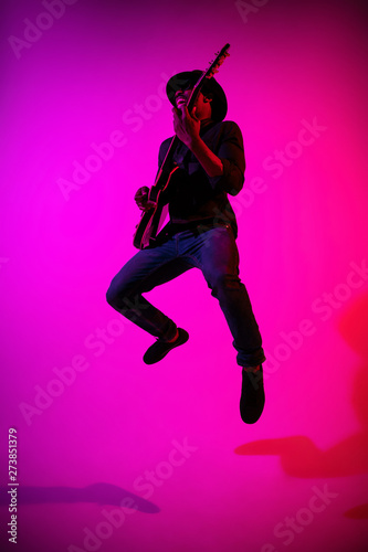 Young african-american musician playing the guitar like a rockstar on gradient purple-pink background in neon light. Concept of music  hobby. Joyful attractive guy improvising. Colorful portrait.