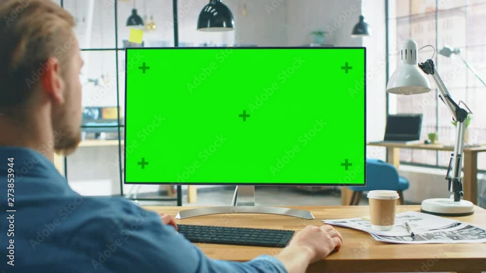 Over the Shoulder Shot: Man Uses Laptop Computer with Green Mock-up Screen while Sitting at His Desk. In the Background Stylish Modern Open Space Creative Business Office
