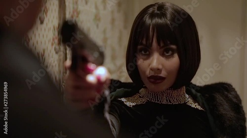 Sexy brunette Arab woman in tight black dress with jewels and big pearl necklace pointing a gun on her husband who cheated on her in a retro hotel with walls adorned with floral tapestry. Close Up.