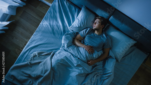 Top View of Handsome Young Man Sleeping Cozily on a Bed in His Bedroom at Night. Blue Nightly Colors with Cold Weak Lamppost Light Shining Through the Window.