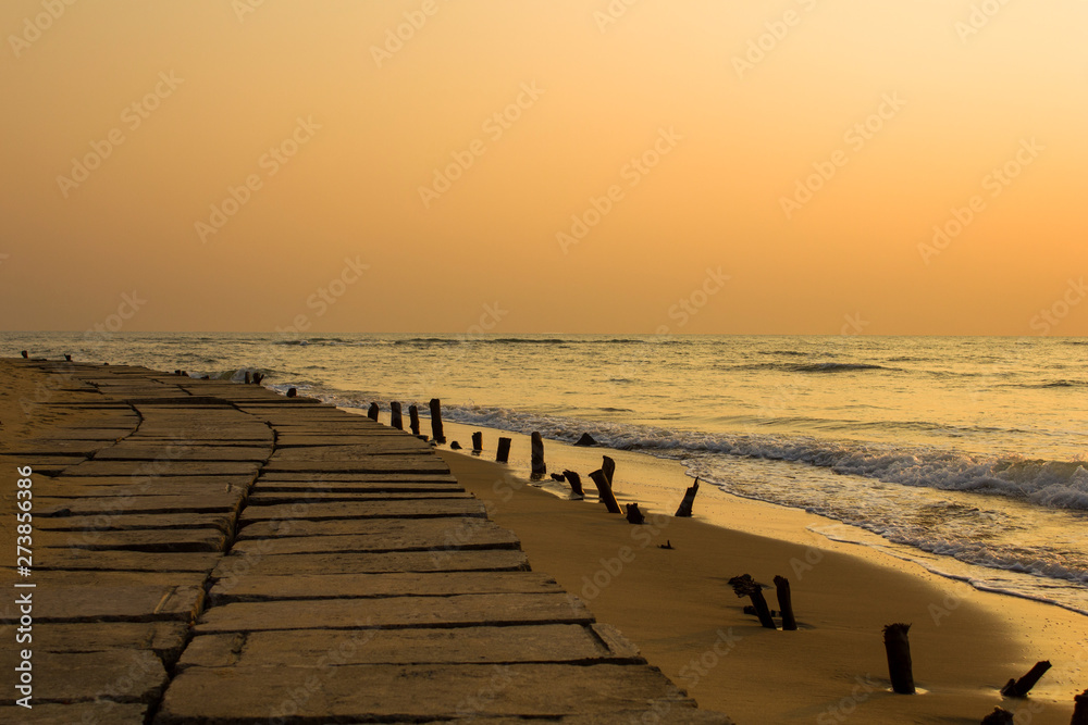 concrete embankment with old wooden columns sticking out of the yellow sand against the background of the ocean waves under a bright orange sunset sky