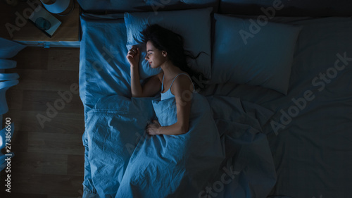 Top View of Beautiful Young Woman Sleeping Cozily on a Bed in His Bedroom at Night. Blue Nightly Colors with Cold Weak Lamppost Light Shining Through the Window. photo