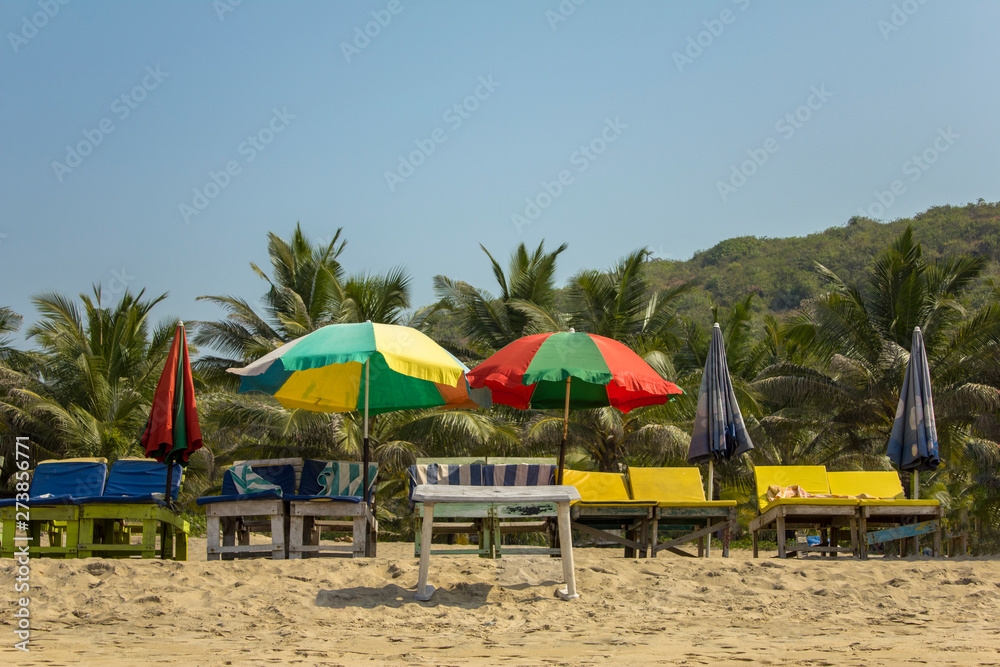 empty sun loungers under the multi-colored beach umbrellas on the yellow sand against the green palm trees under a clear blue sky