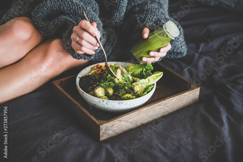 Healthy dinner, lunch setting. Vegan superbowl or Buddha bowl with hummus, vegetable, salad, beans, couscous and avocado, smoothie on tray and woman in warm sweater eating in bed photo