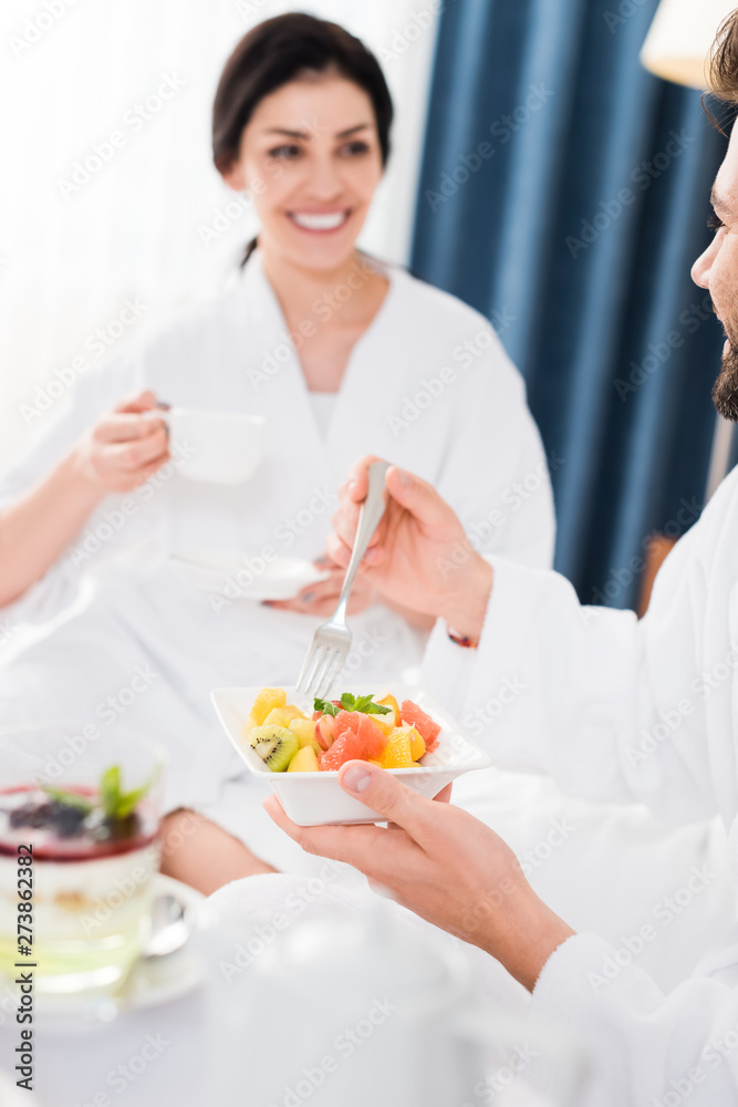 cropped view of cheerful man holding fork near fruit salad and looking at happy woman cropped view of cheerful man holding fork near fruit salad and looking at happy woman