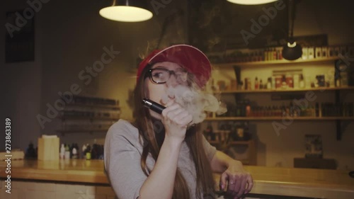 Young pretty woman in red cap smoke an electronic cigarette at the vape shop. Closeup. Slow motion. photo