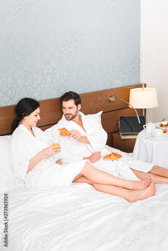 bearded man holding croissant near happy woman with glass of orange juice