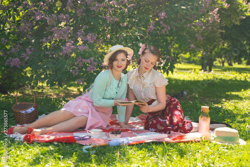 two pretty girls friends sitting on the red blanket on the green grass and have summer picnic. happy woman having rest and fun on their vacantions on the nature. photo