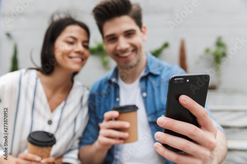 Blurred image of smiling couple drinking takeaway coffee and using smartphone on city stairs outdoors
