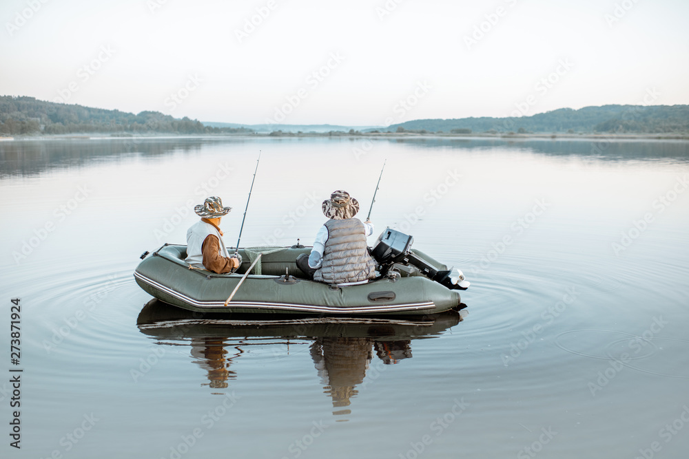 Grandfather with adult son fishing on the inflatable boat on the lake with calm water early in the morning. Wide landscape view
