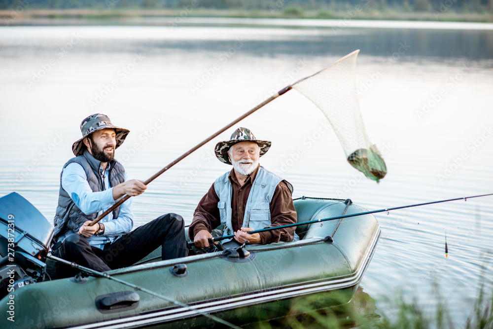 Naklejka premium Grandfather with adult son fishing on the inflatable boat, catching fish with net on the lake early in the morning