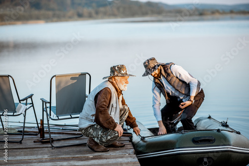 Grandfather with adult son preparing for the fishing, getting on the boat near the pier on the lake early in the morning