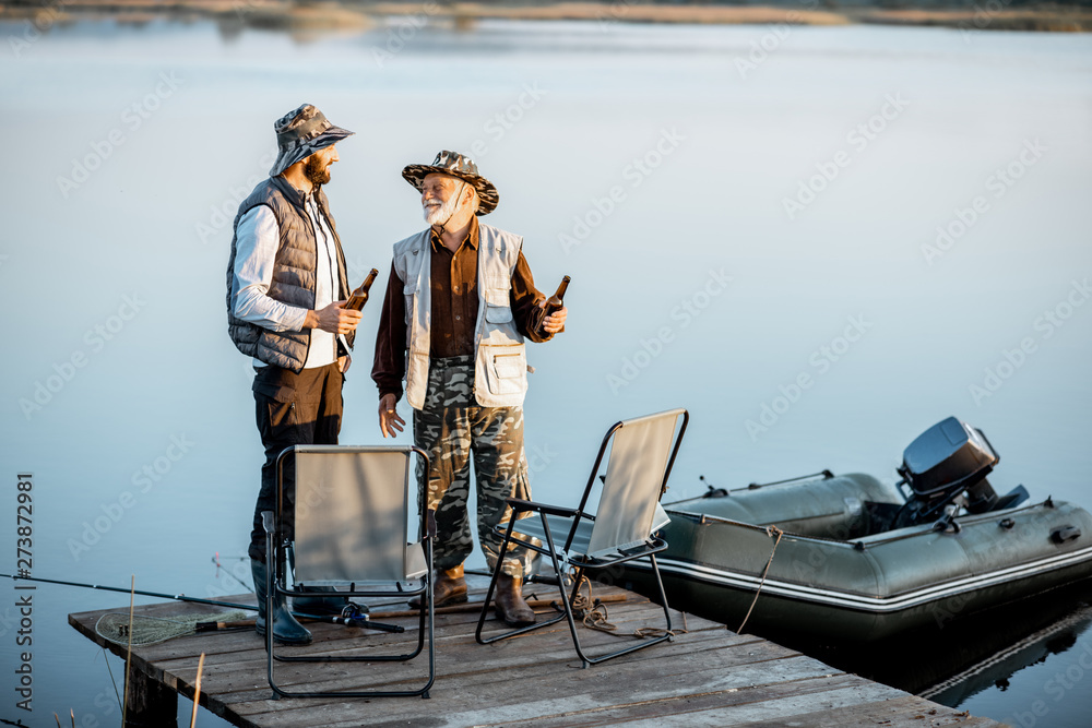 Obraz premium Grandfather with adult son having warm conversation while fishing together on the pier near the lake early in the morning