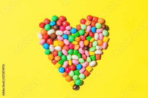 Heart shape made of small colorfull candies on bright yellow background. Sweets for birthday concept. Valentine's day design. Top view, flat lay.
