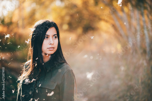 Dreamy beautiful girl in white fluff on bokeh background with yellow leaves in golden sunlight. Inspired girl enjoys sunset in autumn forest. Female beauty portrait among flying spores of thistle.