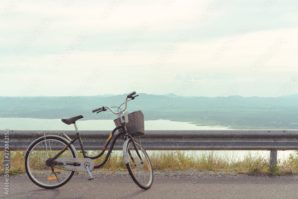 Stationary bicycle on cycle path with amazing scenic views of a lake & mountains - Bike with basket parked next to a cliff edge with epic landscape scenery & warm summer filter - image
