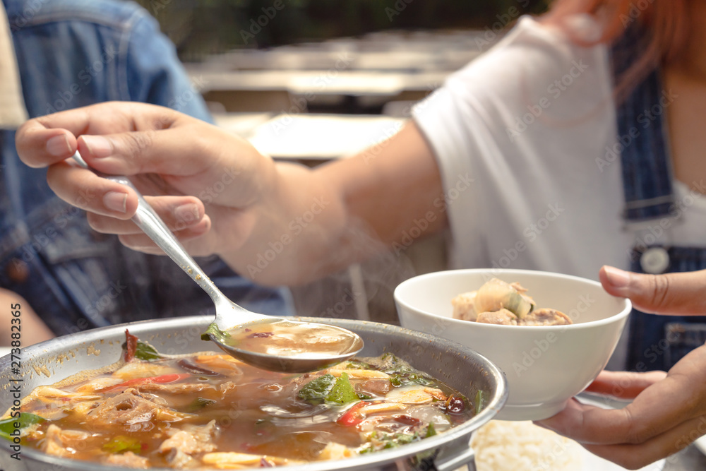 Woman serving steaming hot Asian soup into a bowl at a restaurant during a family meal - Young female diner at a table holding spoon full of Thai Tom Yum spicy soup with summer filter