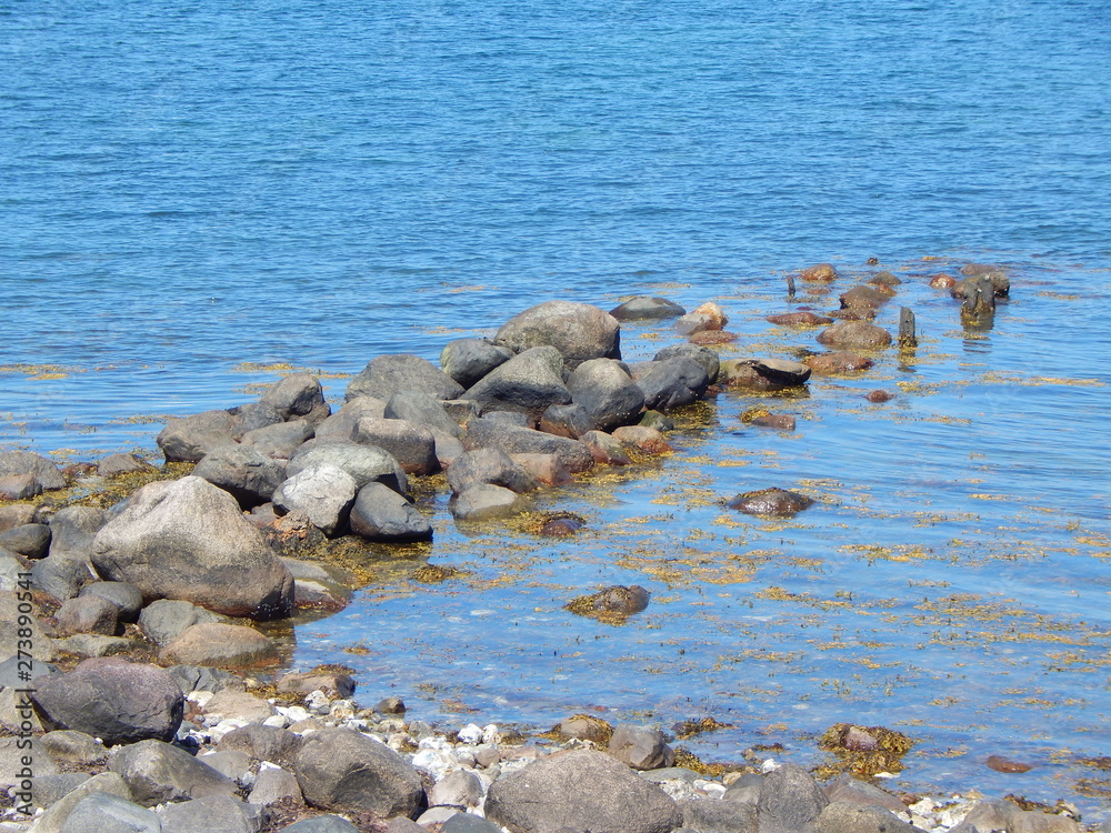 Verfallener Wellenbrecher aus Steinen an der Küste, blaues Wasser und Algen Verfallener Wellenbrecher aus Steinen an der Küste, blaues Wasser und Algen