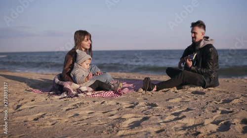 Portrait of a family sits on a plaid blanket on the beach as the father plays the guitar and mother with child listening to him. Cool weather, seasisde. Slow motion photo