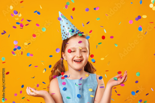 Cheerful little girl celebrates birthday. The child is standing with eyes closed in the rain of confetti. Closeup portrait on yellow background.