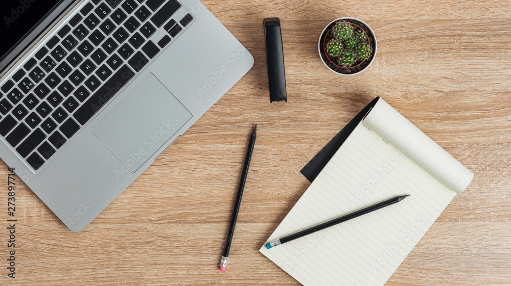 On the desktop, a laptop and a stapler, an open notebook and pencils, and a cactus. View from above. On the desktop, a laptop and a stapler, an open notebook and pencils, and a cactus. View from above.