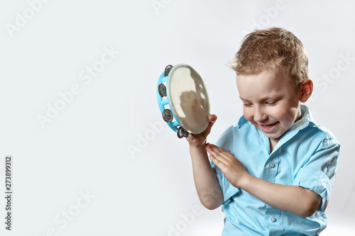 Fototapeta a cheerful boy in a blue shirt holding a tambourine and smiling on a light backg