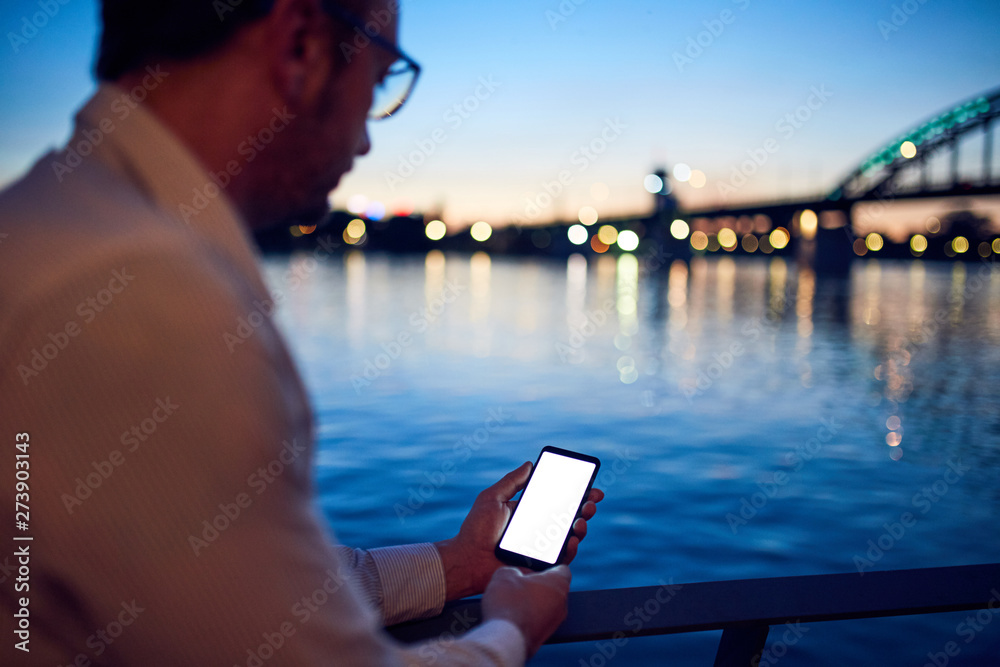 Businessman using cellphone near the river with city lights in background.