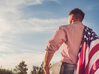 Young engineer, holding white hardhat and an American Flag in the park against the backdrop of green trees and the setting sun, looking into the distance. Close-up. Concept of labor and employment