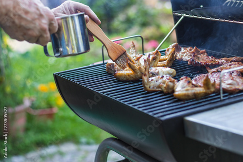 Hand with basting brush marinate meat on grill during garden party. Summer barbecue outdoors