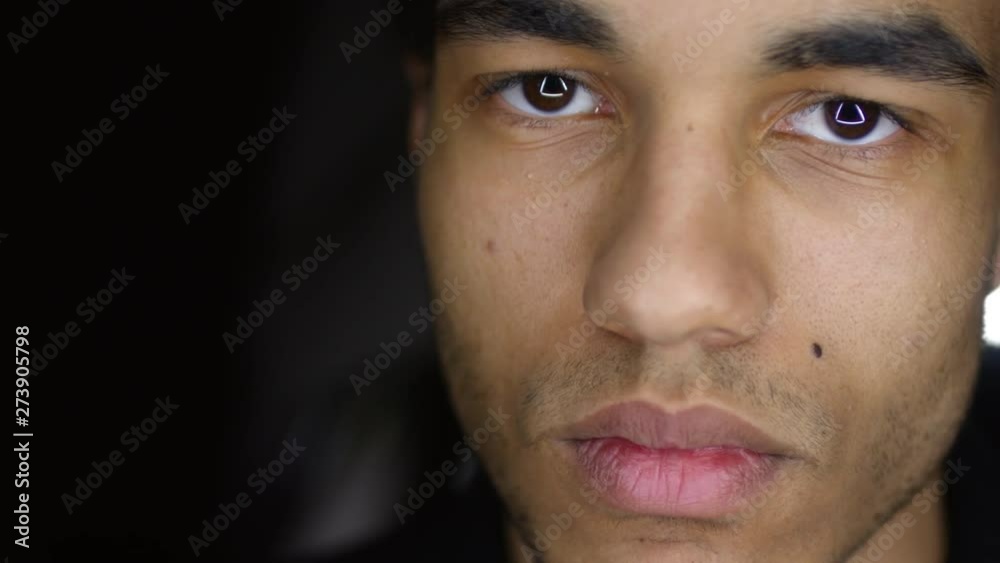 Close up of young mixed-race man looking down then raising his eyes and looking at camera with serious facial expression on black background with neon lamps Close up of young mixed-race man looking down then raising his eyes and looking at camera with serious facial expression on black background with neon lamps