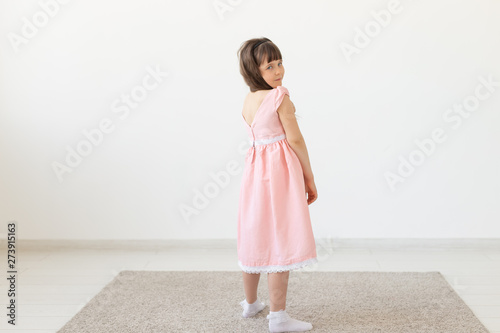 The little charming girl in a pink dress smiles and looks into the camera against the background of a white wall. Concept of the author's children's clothing. Copy space