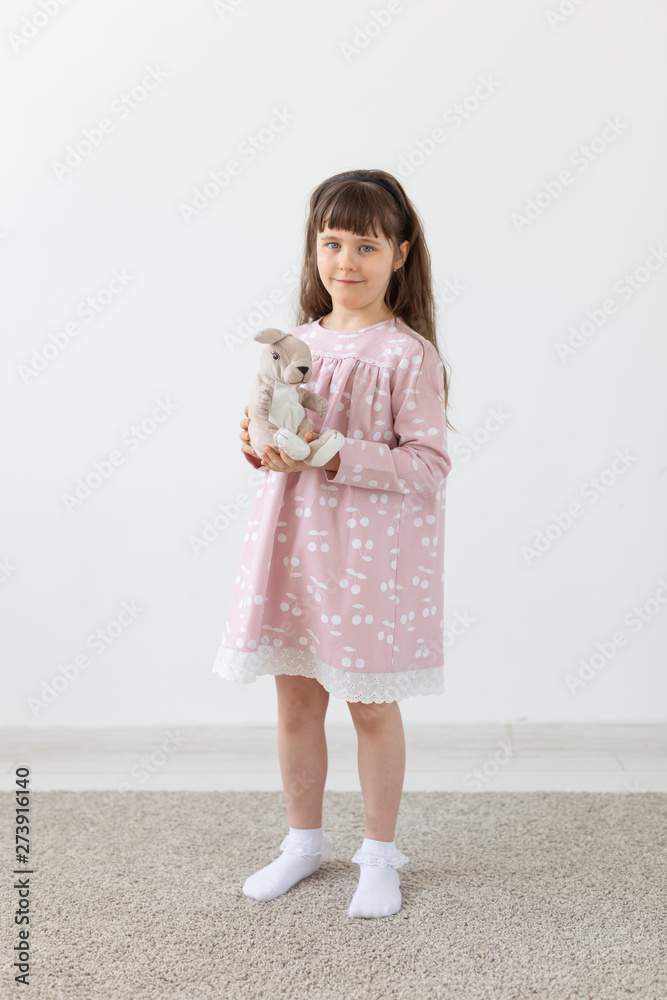 Beautiful little girl holding a teddy rabbit in a pink dress next to a white wall. The concept of cute children and children's toys and clothing. Beautiful little girl holding a teddy rabbit in a pink dress next to a white wall. The concept of cute children and children's toys and clothing.