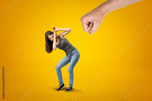 Young brunette girl wearing casual jeans and t-shirt protecting herself from big male stretched fist on yellow background